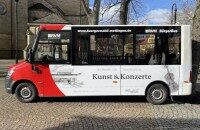 Bürgermobil Mettingen Ein Bild des Bürgermobil Mettingen. Seitenansicht. Der Kleinbus hat große Fenster und ist Weiß mit etwas Rot lackiert.