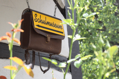 Mettinger Schulmuseum Eine alte Schultasche hängt an einer Hauswand. An ihr ist ein gelbes Schild mit der Aufschrift